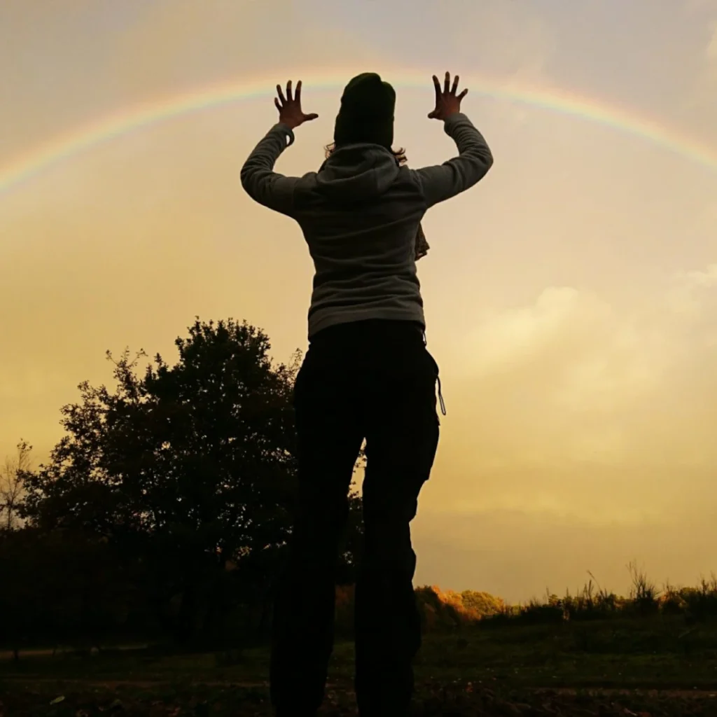 Frau greift mit nach oben gestreckten Händen nach einem Regenbogen